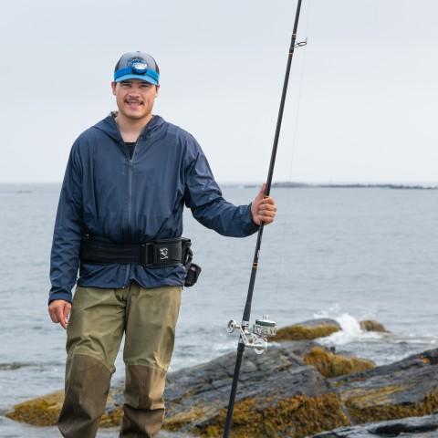 A UNE student poses with a fishing rod in front of the ocean